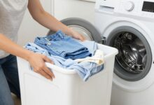 Uma pessoa sorrindo enquanto organiza roupas coloridas e brancas em cestos de lavanderia, com uma máquina de lavar moderna ao fundo, ilustrando a organização para a lavagem de roupas.