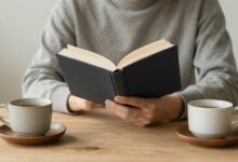 Uma pessoa sentada calmamente em um ambiente minimalista e iluminado, lendo um livro com uma xícara de chá ao lado, transmitindo paz e tranquilidade.