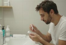 Homem adulto em ambiente claro realizando cuidados diários de higiene e bem-estar, com produtos de pele, água e roupas esportivas ao redor