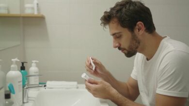 Homem adulto em ambiente claro realizando cuidados diários de higiene e bem-estar, com produtos de pele, água e roupas esportivas ao redor